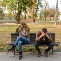 An upset couple seated on a park bench, expressing frustration during an autumn day.